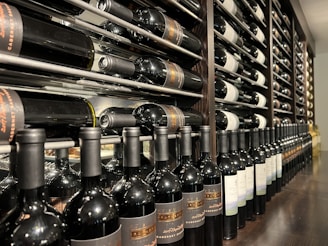 A well-organized wine cellar with rows of wine bottles stored horizontally on wooden racks. The bottles have elegant labels, indicating different varieties and vintages. The setting is dimly lit, suggesting a sophisticated, serene atmosphere typically associated with wine storage.