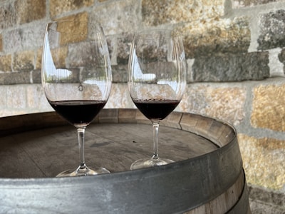 Two wine glasses with red wine placed on top of a wooden wine barrel. The background consists of a rustic stone wall with various shades of brown, gray, and tan.