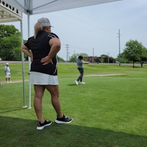 A person wearing a skirt and black shirt stands under a canopy, observing another individual who is swinging a golf club on a green course. Another person in casual attire is visible in the background, watching. The scene is outdoors with a backdrop of trees and power lines.