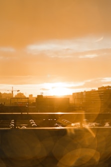 The image captures a city skyline during sunset, with the sun descending near the horizon, casting a warm orange and golden hue throughout the scene. Tall buildings and cranes are silhouetted against the vibrant sky, and lens flares add an artistic element to the composition.