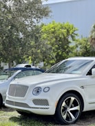 A white luxury SUV is parked outdoors near trees. The vehicle features a distinctive front grille and large wheels. Another car is partially visible in the background, with lush greenery surrounding the scene.
