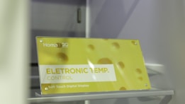 A sign is displayed with yellow and white colors featuring the text 'ELETRONIC TEMP. CONTROL' along with 'Soft Touch Digital Display.' The background has a soft yellow hue with circular patterns resembling bubbles or holes. The brand 'Homa' is visible along with a 20th-anniversary symbol.