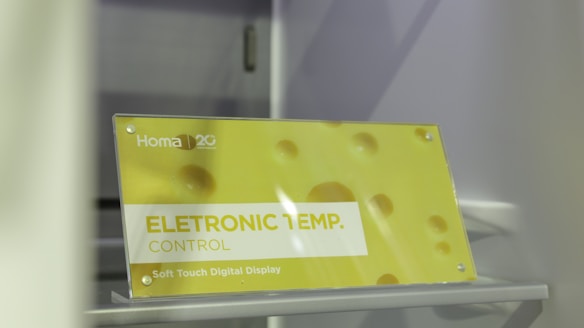 A sign is displayed with yellow and white colors featuring the text 'ELETRONIC TEMP. CONTROL' along with 'Soft Touch Digital Display.' The background has a soft yellow hue with circular patterns resembling bubbles or holes. The brand 'Homa' is visible along with a 20th-anniversary symbol.