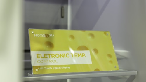 A sign is displayed with yellow and white colors featuring the text 'ELETRONIC TEMP. CONTROL' along with 'Soft Touch Digital Display.' The background has a soft yellow hue with circular patterns resembling bubbles or holes. The brand 'Homa' is visible along with a 20th-anniversary symbol.