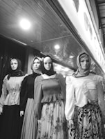 Four mannequins dressed in a variety of long-sleeved blouses and flowing skirts, each wearing a headscarf. The mannequins are displayed in a storefront with a reflective ceiling creating a pattern above. The image is in black and white, emphasizing contrast and texture.