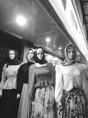 Four mannequins dressed in a variety of long-sleeved blouses and flowing skirts, each wearing a headscarf. The mannequins are displayed in a storefront with a reflective ceiling creating a pattern above. The image is in black and white, emphasizing contrast and texture.