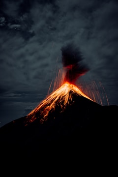 A volcano actively erupting with bright lava spewing from the summit, creating a striking contrast against the dark, cloudy sky. The eruption casts an intense glow, illuminating the ash plume rising into the air.