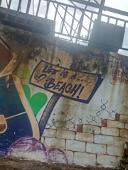 The image features wall art depicting a signpost with the phrase 'Life is a... Beach!' in bold letters. The background includes a textured, weathered surface with bricks and decorative elements like a heart shape and a stylized vine or fence. The overall aesthetic suggests a rustic or graffiti-style mural.