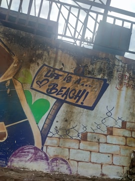 The image features wall art depicting a signpost with the phrase 'Life is a... Beach!' in bold letters. The background includes a textured, weathered surface with bricks and decorative elements like a heart shape and a stylized vine or fence. The overall aesthetic suggests a rustic or graffiti-style mural.