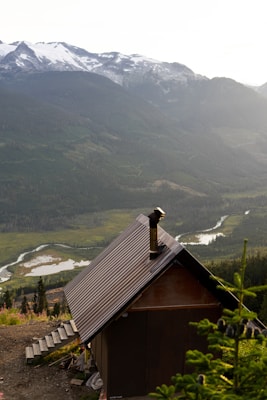 A rustic cabin with a metal roof is situated on a hillside, overlooking a vast landscape. The background features expansive mountains with snow-capped peaks and lush green forests. A winding river can be seen snaking through the valley below, and the overall atmosphere is peaceful and serene.