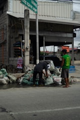 Two individuals are interacting with several bags of garbage on the side of a street, in front of a building with an open garage and a sign above. The setting appears to be urban, with visible infrastructure including a pole and wires.