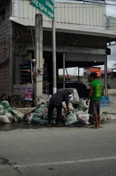 Two individuals are interacting with several bags of garbage on the side of a street, in front of a building with an open garage and a sign above. The setting appears to be urban, with visible infrastructure including a pole and wires.