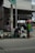 Two individuals are interacting with several bags of garbage on the side of a street, in front of a building with an open garage and a sign above. The setting appears to be urban, with visible infrastructure including a pole and wires.