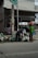 Two individuals are interacting with several bags of garbage on the side of a street, in front of a building with an open garage and a sign above. The setting appears to be urban, with visible infrastructure including a pole and wires.