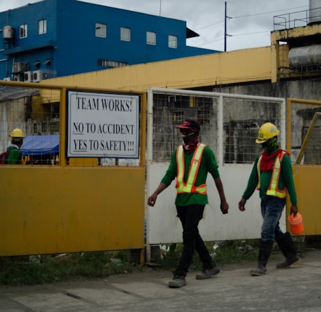 Two construction workers wearing safety vests and helmets walk past a construction site. A sign on the fence reads, 'TEAM WORKS! NO TO ACCIDENT YES TO SAFETY!!!' The background includes a building with blue and yellow exterior colors.