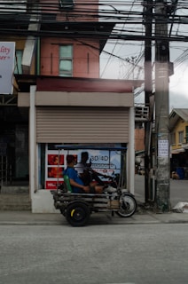 A tricycle vehicle with two passengers is parked on the side of a street. The vehicle appears to be makeshift, with a metal frame and a simple seating arrangement. Behind the tricycle is a closed storefront with advertisements, including one for ultrasound services. Overhead, numerous electrical wires are visible.