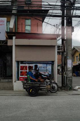 A tricycle vehicle with two passengers is parked on the side of a street. The vehicle appears to be makeshift, with a metal frame and a simple seating arrangement. Behind the tricycle is a closed storefront with advertisements, including one for ultrasound services. Overhead, numerous electrical wires are visible.