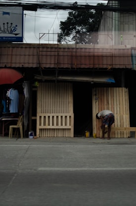 A small roadside shop with wooden pallets leaning against its entrance. There's a person bending over near the pallets, possibly working or picking something up. An umbrella with clothes hanging and a plastic chair is to the left of the shop. Electrical wires and a tree are visible above the shop.