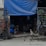 A makeshift shelter with a blue tarp covering. Inside, a person is sitting on a chair, while another is lying in a hammock. A small dog sits alertly at the entrance. Various tools and cables are visible, and small potted plants are arranged on shelving.