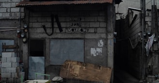 A small, weathered building made of concrete blocks with a corrugated metal roof. The facade displays a sign with the words 'Mayora Jon We Love You'. To the left, there are stacked cinder blocks and wooden planks. A narrow alley is visible on the right side, with numerous exposed wires and electrical meters attached to the wall. The overall atmosphere is somewhat rundown and urban.