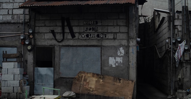 A small, weathered building made of concrete blocks with a corrugated metal roof. The facade displays a sign with the words 'Mayora Jon We Love You'. To the left, there are stacked cinder blocks and wooden planks. A narrow alley is visible on the right side, with numerous exposed wires and electrical meters attached to the wall. The overall atmosphere is somewhat rundown and urban.