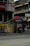 A sidewalk scene featuring a small street vendor setup with a colorful umbrella, containing various goods on display. A person is standing next to the stall. Nearby buildings have signs, some partially visible, advertising local businesses. The architecture is a mix of modern and older structures.