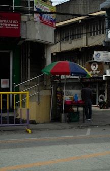 A sidewalk scene featuring a small street vendor setup with a colorful umbrella, containing various goods on display. A person is standing next to the stall. Nearby buildings have signs, some partially visible, advertising local businesses. The architecture is a mix of modern and older structures.