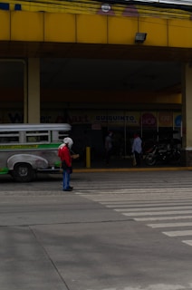 A person wearing a red shirt and blue pants stands in front of a pedestrian crossing on an urban street. The background shows a jeepney parked nearby and a few people standing near a commercial establishment with signs visible. The building has a prominent yellow awning and there is a motorcycle parked in front.