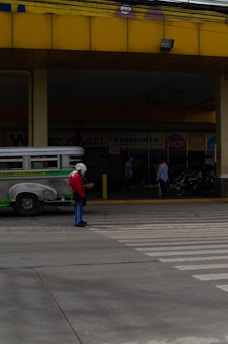 A person wearing a red shirt and blue pants stands in front of a pedestrian crossing on an urban street. The background shows a jeepney parked nearby and a few people standing near a commercial establishment with signs visible. The building has a prominent yellow awning and there is a motorcycle parked in front.