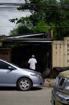 A person wearing a white shirt and hat is standing near the entrance of a garage or workshop. In front of the entrance, there are several parked vehicles, including a silver car in the foreground. The garage seems cluttered with items including tires. Overhead, there are several power lines, and the area is surrounded by greenery.