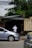 A person wearing a white shirt and hat is standing near the entrance of a garage or workshop. In front of the entrance, there are several parked vehicles, including a silver car in the foreground. The garage seems cluttered with items including tires. Overhead, there are several power lines, and the area is surrounded by greenery.
