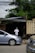 A person wearing a white shirt and hat is standing near the entrance of a garage or workshop. In front of the entrance, there are several parked vehicles, including a silver car in the foreground. The garage seems cluttered with items including tires. Overhead, there are several power lines, and the area is surrounded by greenery.