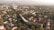 An aerial view of a cityscape with a mix of residential and commercial buildings surrounded by abundant greenery. In the background, mountains are visible under a cloudy sky. The residential area features houses with colorful roofs, while taller buildings can be seen in the distance.
