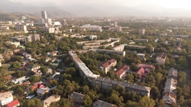 An aerial view of a cityscape with a mix of residential and commercial buildings surrounded by abundant greenery. In the background, mountains are visible under a cloudy sky. The residential area features houses with colorful roofs, while taller buildings can be seen in the distance.