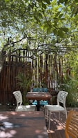 A serene outdoor seating area featuring a wooden table surrounded by four plastic chairs. Overhead, a lush canopy of green leaves provides shade, while rustic bamboo fencing adds to the natural ambiance. String lights are hung among the branches, suggesting a cozy atmosphere in the evening.