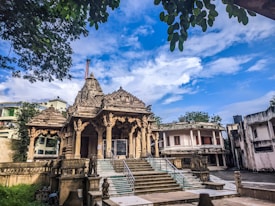 An intricately designed temple with ornate carvings and pillars, surrounded by a few residential buildings. The temple has a series of steps leading up to it and is flanked by statues. Above, the sky is bright with scattered clouds and leaves from nearby trees frame the image.