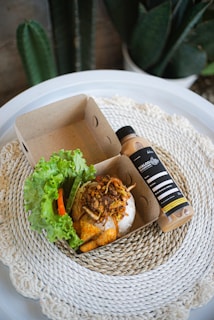 A small cardboard food container holds a serving of seasoned rice topped with fried shallots and meat, accompanied by fresh lettuce, cucumber, and chili. Alongside it is a bottle of a beverage labeled 'DIALOGUE' placed on a round, woven placemat. The background includes green houseplants.