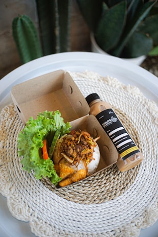 A small cardboard food container holds a serving of seasoned rice topped with fried shallots and meat, accompanied by fresh lettuce, cucumber, and chili. Alongside it is a bottle of a beverage labeled 'DIALOGUE' placed on a round, woven placemat. The background includes green houseplants.