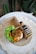 A small cardboard food container holds a serving of seasoned rice topped with fried shallots and meat, accompanied by fresh lettuce, cucumber, and chili. Alongside it is a bottle of a beverage labeled 'DIALOGUE' placed on a round, woven placemat. The background includes green houseplants.