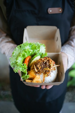 A person is holding a container filled with a variety of foods which includes fresh green lettuce, tomato, cucumber slices, rice, and a mixture of fried or sautéed elements. The person is wearing a dark apron over a light-colored, patterned long-sleeve shirt.