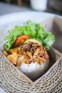 A portion of rice topped with a variety of seasoned ingredients, including crispy onions and bits of meat, arranged in a cardboard container. Fresh green lettuce and sliced tomatoes are set beside the rice, accompanied by some crispy crackers. The box rests on a woven placemat, giving it a rustic and appetizing presentation.