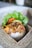 A portion of rice topped with a variety of seasoned ingredients, including crispy onions and bits of meat, arranged in a cardboard container. Fresh green lettuce and sliced tomatoes are set beside the rice, accompanied by some crispy crackers. The box rests on a woven placemat, giving it a rustic and appetizing presentation.