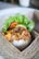 A portion of rice topped with a variety of seasoned ingredients, including crispy onions and bits of meat, arranged in a cardboard container. Fresh green lettuce and sliced tomatoes are set beside the rice, accompanied by some crispy crackers. The box rests on a woven placemat, giving it a rustic and appetizing presentation.