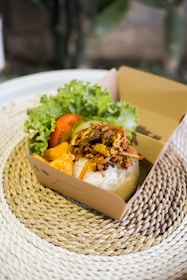 A cardboard box contains a serving of white rice topped with seasoned shredded meat. Accompanying the rice inside the box are slices of fresh lettuce, tomato, and a piece of deep-fried orange-colored snack. The box is placed on a textured woven placemat, providing a rustic feel.