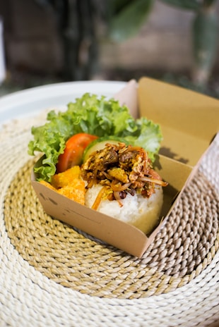 A cardboard box contains a serving of white rice topped with seasoned shredded meat. Accompanying the rice inside the box are slices of fresh lettuce, tomato, and a piece of deep-fried orange-colored snack. The box is placed on a textured woven placemat, providing a rustic feel.