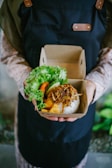 A person wearing a dark apron holds a cardboard container with a meal. The container includes rice topped with crispy fried onions, tofu, and a side of fresh greens such as lettuce and slices of cucumber and tomato.