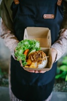 A person wearing a dark apron holds a cardboard container with a meal. The container includes rice topped with crispy fried onions, tofu, and a side of fresh greens such as lettuce and slices of cucumber and tomato.
