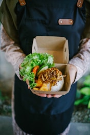 A person wearing a dark apron holds a cardboard container with a meal. The container includes rice topped with crispy fried onions, tofu, and a side of fresh greens such as lettuce and slices of cucumber and tomato.