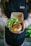 A person wearing a dark apron holds a cardboard container with a meal. The container includes rice topped with crispy fried onions, tofu, and a side of fresh greens such as lettuce and slices of cucumber and tomato.