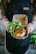 A person wearing a dark apron holds a cardboard container with a meal. The container includes rice topped with crispy fried onions, tofu, and a side of fresh greens such as lettuce and slices of cucumber and tomato.